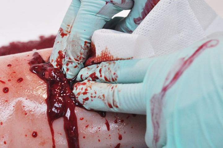Close-up of gloved hands applying TrueClot simulated gauze into a moulage wound with artificial blood during trauma training.
