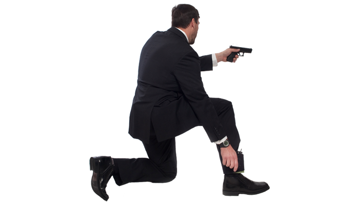 Person in tactical attire kneeling with weapon drawn, wearing ankle medical kit under trousers.