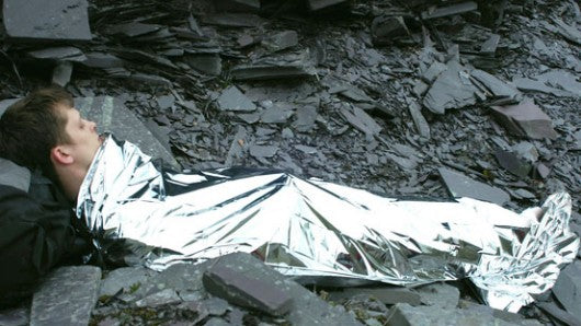 Person lying outdoors wrapped in a reflective emergency thermal blanket.