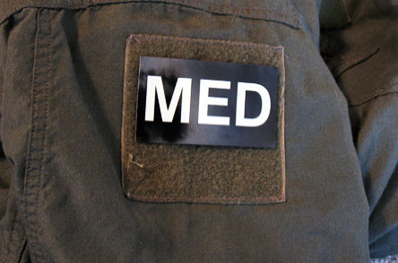 Glow-in-the-dark 'MED' patch affixed to worn outdoor jacket, shown in field conditions.