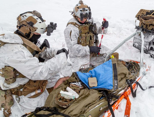 Close-up of HPMK-I™ (Insulated) being deployed in a snowy field by two tactical medics in white gear. The tan and blue blanket is opened, showcasing its use for thermal protection in harsh conditions.